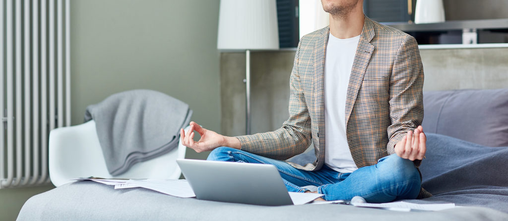 Full length portrait of handsome young freelancer meditating in lotus position while taking break from work, documents and laptop located on cozy bed, interior of studio apartment on background
